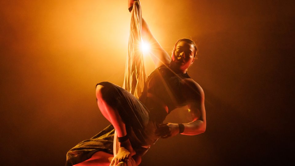 A woman with dark hair pulled back has her right arm raised in the air to hold onto a dangling silk. Her legs are crouched with her feet perched onto the dangling silk. She is lit from behind with an orange glow.