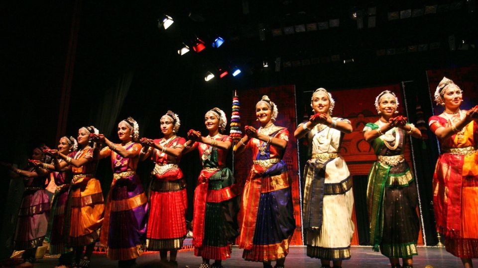 A group of female Indian dancers are lined up across a stage proscenium. They are all wearing traditional Indian dance clothes.