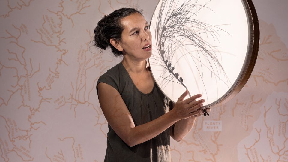 A woman with brown hair tied up in a bun is wearing a brown short sleeved top. She holds a large drum in her left hand slung on top of her shoulder. In her right hand is a bush feather.