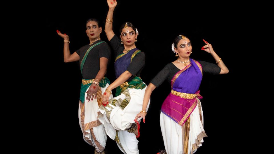 Three Indian women wearing white balloon pants and purple tops, stand next to each other with their arms raised in the air and feet dancing.