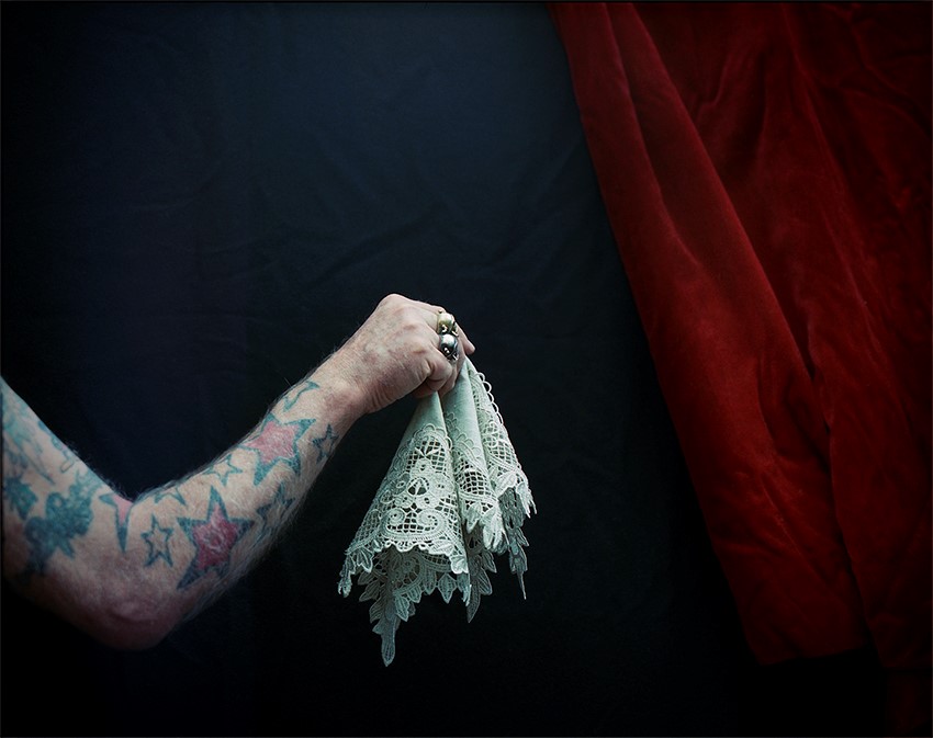 Image of a tattoo arm with hand with rings on holding an old lace hankerchief in the foreground.  In the background red velvet curtains. 
