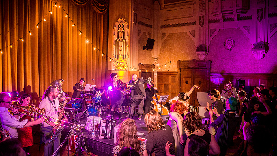 A crowd of people dancing and playing music in a hall with orange and pink light reflecting off the walls