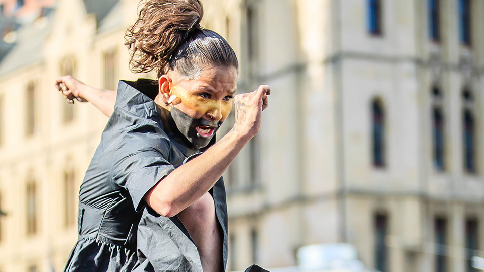 A person wearing face-paint and a dark ponytail in focus in mid-leap, with a blurry Melbourne street in the background