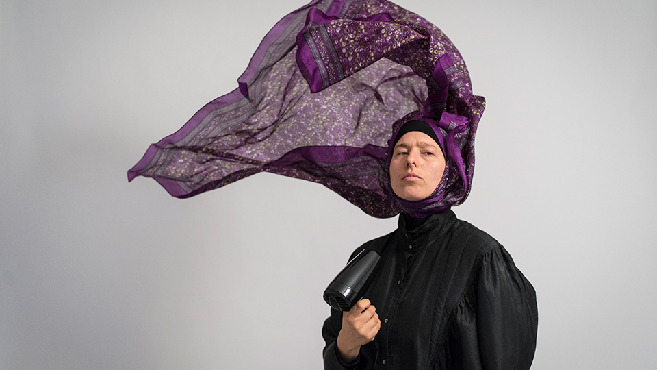 Person with hairdryer pointed at their face, making their purple headscarf lift into the air