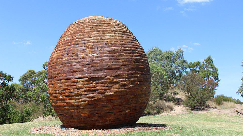A large brown stone nest sculpture sitting in a park with trees in the background. 