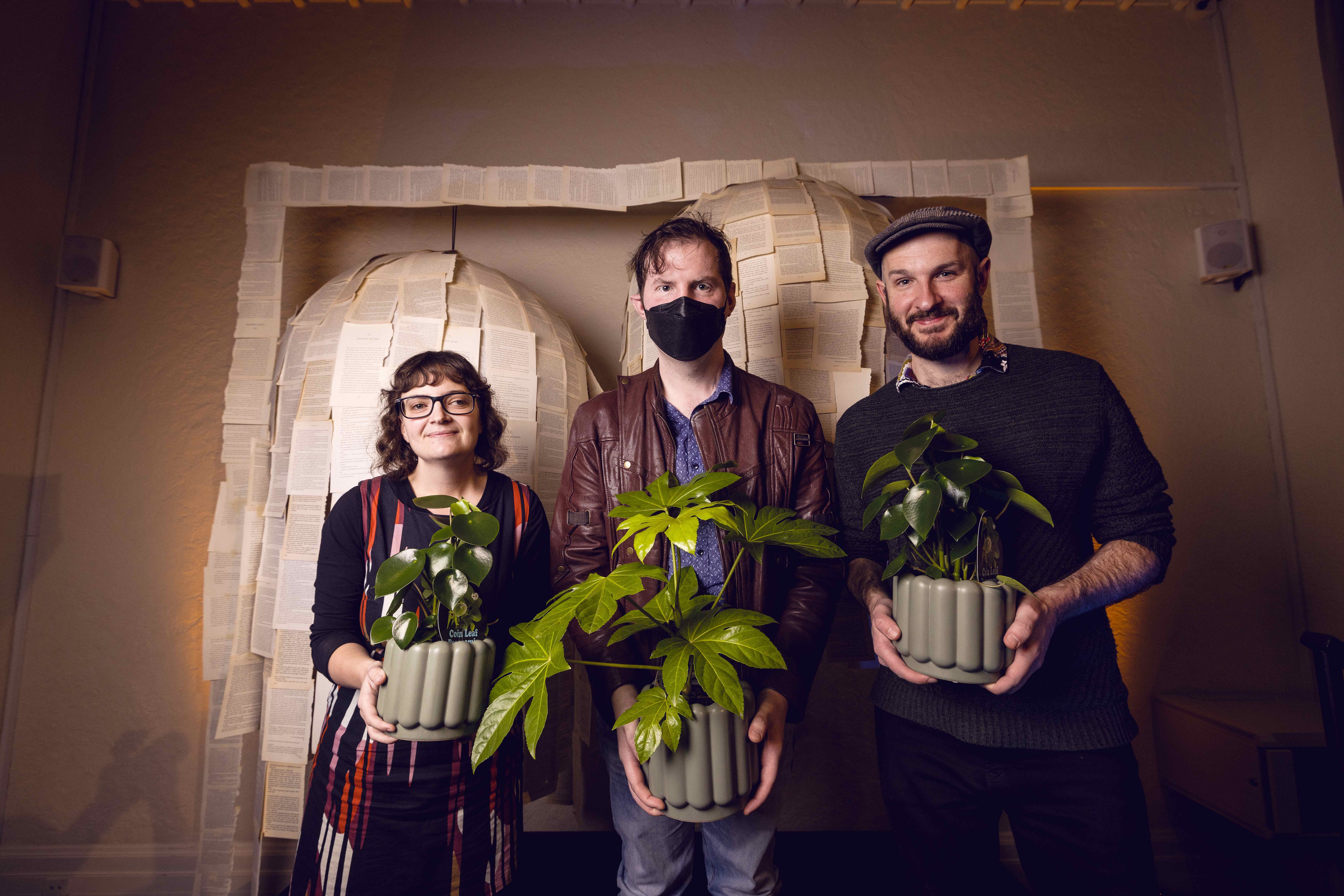 A woman wearing glasses and a long sleeve dark coloured dress, and two men: one wearing a balck mask over his mouth and the other wearing a brown golfer's hat, stand side by side in front a beige wall with a literary page structure. All three people are holding green plants in front of them.