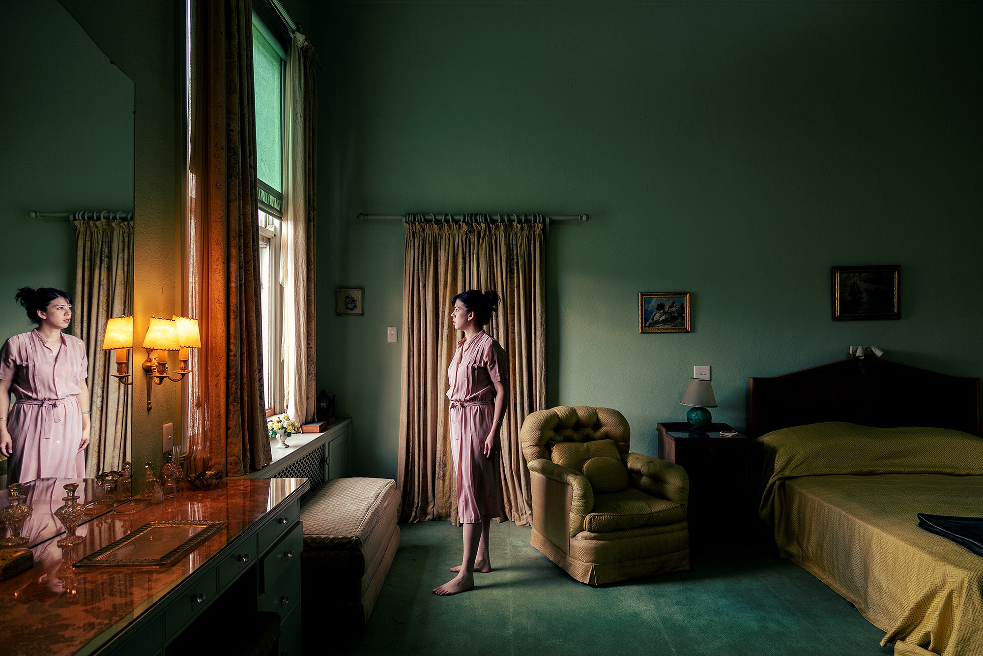 Pia Johnson's artwork 'A view of the pool' from Mooramong Green series, 2020, Archival inkjet print. A woman stands before an open bedroom window and gazes out, her reflection appears in a mirror in the foreground of the image. 