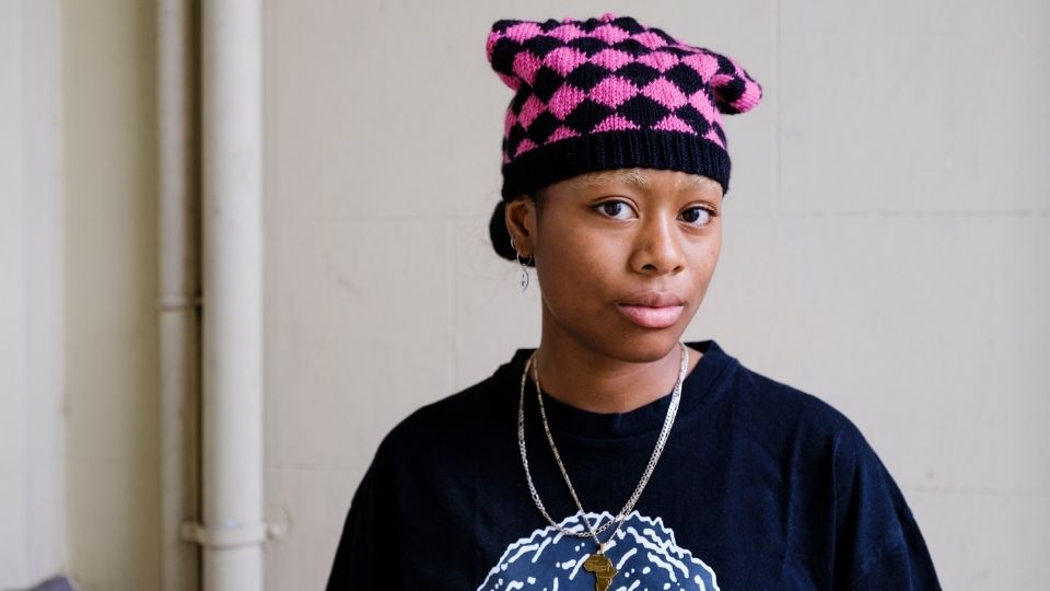 A young black woman wearing a pink and black checker beanie hat, a black t-shirt and two silver metal necklaces smiles at the camera facing left and turning her head right slightly.