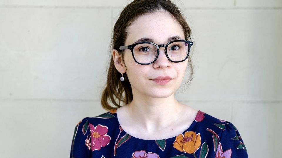 A young woman wearing dark framed glasses has her brown hair pulled back. She is wearing a navy blue floral shirt.