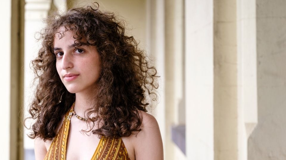 A young woman with medium length, curly hair stands in slight profile to smile to the camera. She is wearing a mustard yellow tank top and a beaded necklace.
