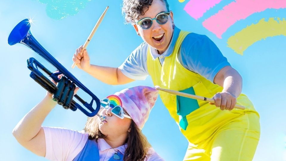 Two people wearing bright coloured overalls and t-shirts pose with instruments: one person with long brown hair and blue framed glasses holds a blue trumpet to their lips. Another person stands to the right of the other person with drum sticks in each hand. There is a small rainbow in a blue sky to the right of the people.