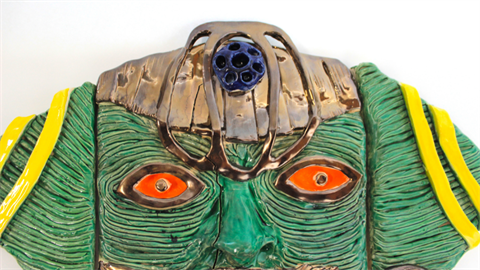 A green ceramic face with orange eyes and a gold head adornment 