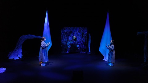 Two women standing at both sides of a lit stage wearing long garments. The stage is lit in a blue hue.
