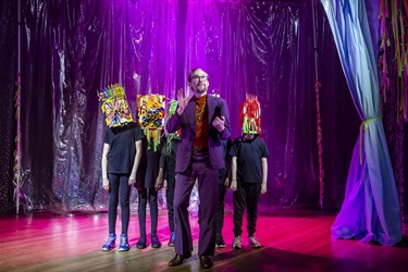 A man in a brown suit stands awkwardly in front of a group of young people wearing boxes on over their faces.