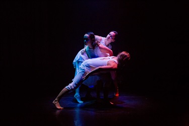 Three female dancers wear white outfits and crouch around and over each other in the centre of a dark stage.