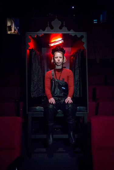 A person with short light hair sits in an oversized armchair with red glowing behind them.
