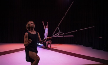 A man wearing a black tank top and black shorts lunges on his left knee. He holds a camera tripod in his right hand. A person wearing a white outfit stands in the background with arms raised in the air.