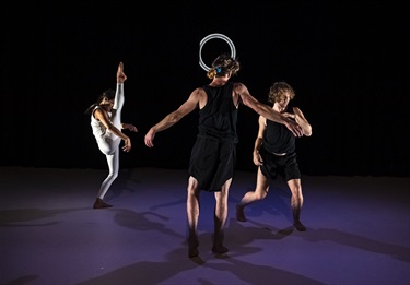 Three people are scattered in various positions on a purple floor. A woman wearing a white outfit stretches her one leg in the air with toes pointed. A thin white circle hovers in the black background.