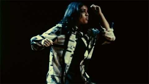 A woman with long dark hair wears a long-sleeves denim jacket and raises her bent left arm towards her forehead.