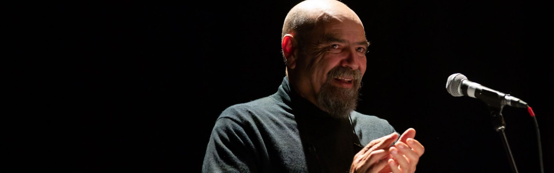 A bald headed man applauds under a spotlight and next to a microphone stand.