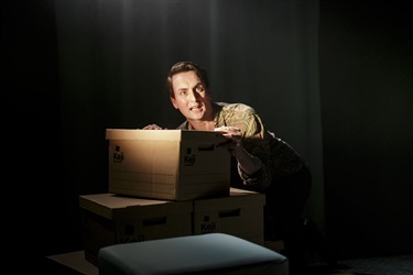 A man peering out from behind a stack of cardboard boxes.