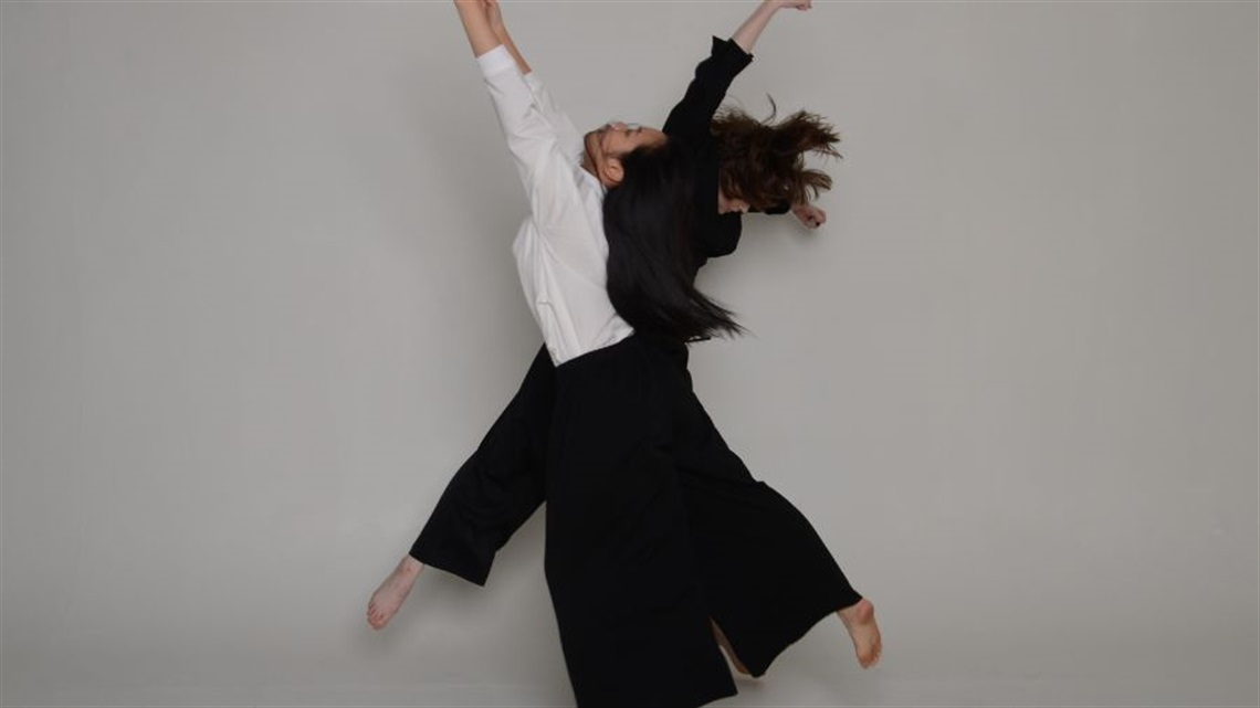 Two female dancers wearing black pants and white tops leap in the air stretching their arms above their heads and pointing their toes.