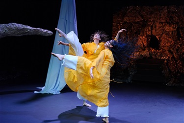 Two female dancers, one with long dark hair, the other with mid-length brown hair, stretch their right legs into the air. They both wear white pants and yellow moomoos.