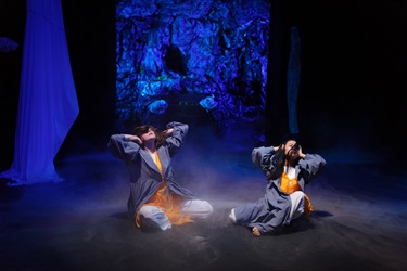 Two women sit on a hazy dark stage. They raise their elbows to the air while holding their hands to their ears and look up to the sky. The background is blue.