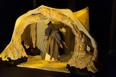 Two people stand on their legs and bend slightly forward. They are inside a large tent-like structure, wearing long dark clothing and white rimmed hats.