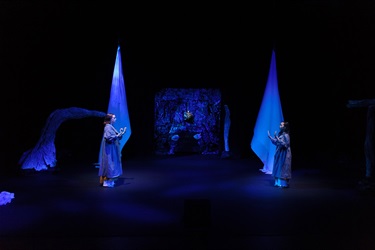 On a dark black stage with flicks of blue hue stand two women on either side of the stage. They face each other and hold their hands towards one another.