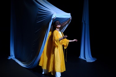 A woman wearing a yellow gown and shite pants stretches her arms out with her elbows closest to her. Someone behind her stretches a white sheet over her like a shady shelter.