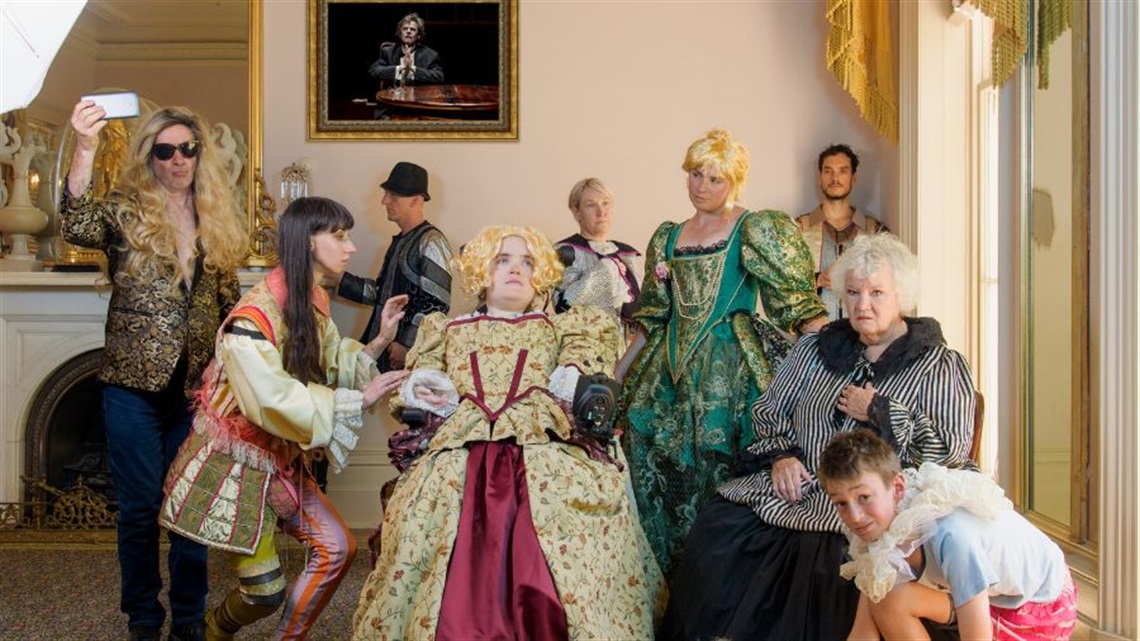 A group of people in a Victorian-period lounge room cluster around a person sitting in a wheelchair. They are all dressed in Victoria-era costumes.