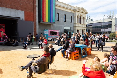 A crowd of people sit and stand in a public square facing a large building with a black stage set up.