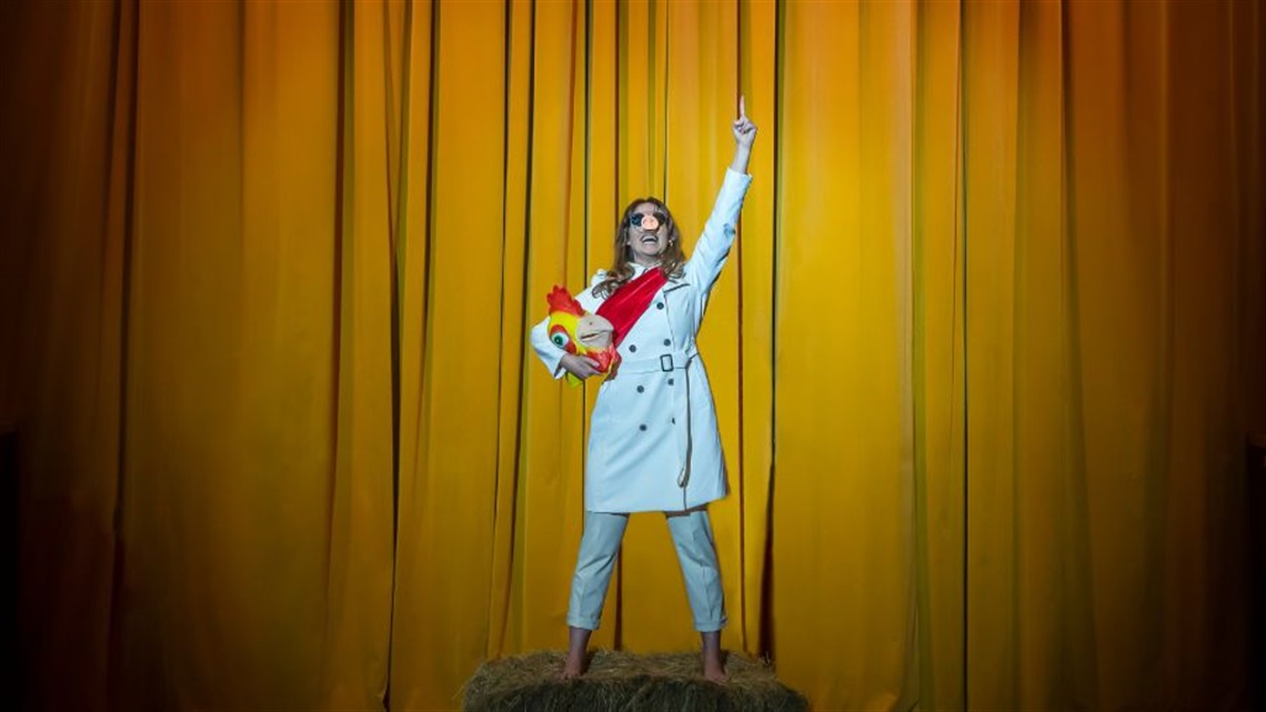 A woman with long hair wears a white trench coat, white pants, sunglasses and a pig nose on her face. She holds a large fake chicken's head on her right arms while her left arm is riased pointing to the sky.