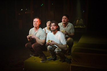 Three men wearing white tshirts and dark pants crouch in a cluster. Their arms are bent with hands clasped together. They look nervously into the distance.