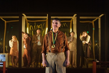 A man with short dark hair and wearing a buttoned shirt with the sleeves rolled up stands at the centre of the stage. A gold medal hangs from his neck. He slyly smiles to the camera.