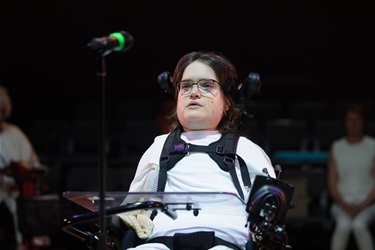 A woman wearing thin-framed glasses and a white t-shirt sits in a wheelchair.