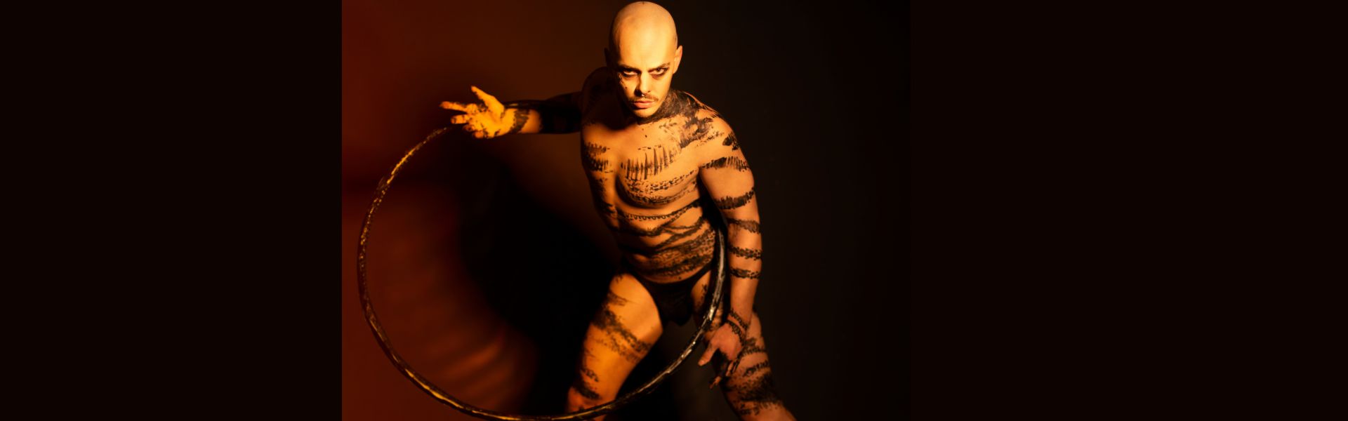 A bald-headed man holds a hula hoop in his criss-crossed hands around his upper body. He stands slightly crouching forward through the centre of the hula hoop. Black marks cover his nearly naked body. 