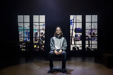 A boy with long dark hair sits centre stage. He wears a striped tshirt underneath a grey hoodie and headphones around his neck. Behind him are two vertical windows frames.