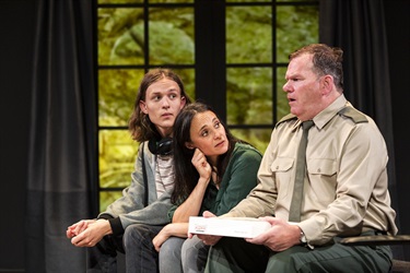 Two men and a woman sit next to each other. One man is dressed like a military officer and holds a thin white box in her hands. The other man has headphones around his neck and long hair. The woman leans forward resting her cheek on her right hand.