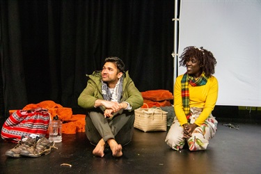 A man with short dark hair and wearing a green jacket sits on a black floor with his knees to his chest and arms around his knees. A woman with short dark curly hair sits on her knees to the man's right. She is also wearing a red scarf around her neck.