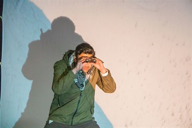 A man with short dark hair stands in a spot light with his hands over his eyes like binoculars.