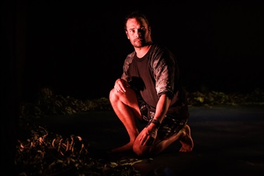 A man with dark hair facial hair and dark hair pulled back into a bun crouches to the ground. He wears a dark tshirt and dark shorts. Gumtree leaves lay beside him on the floor.