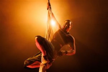 A woman wearing baggy cropped pants holds tight to a silk rope. Her legs are bent so that her feet clutch to the silk rope. A bright yellow light shines behind her.