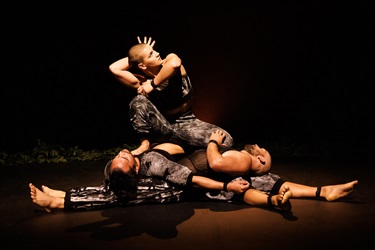 A woman crouches on top of a pile of humans. She has short dark hair and is wearing dark pants. Her right hand is behind her head as she looks over her left shoulder and fans her right hand's fingers.