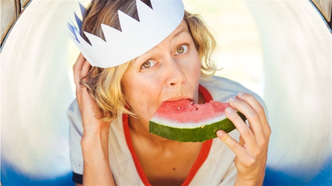 A woman with short light hair wears a white paper crown on her head and a white tshirt with a red neckline. She eats a slice of watermelon.