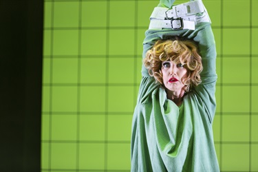 A woman with short blonde curly hair is wearing a green mumu. Her hands are tied above her head and a white cloth is tied around her head blocking her mouth.