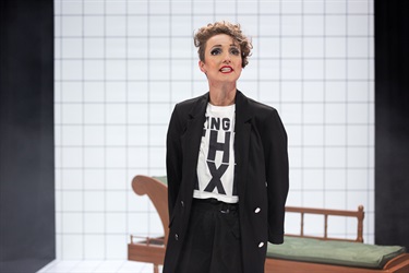 A woman with short light hair and dark eyes wears a black and white tshirt under a black blazer jacket. She stands with confidence as if addressing a crowd.s