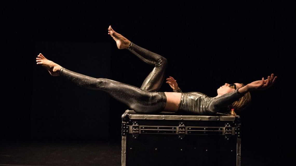 A woman lies on her back on top of a black box. She stretches her legs while pointing her toes, and her arms out to the sides. She is wearing goggles on her eyes and a metallic body suit.