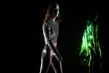 A woman wearing make up on her face and a silver shiny body suit walks forward in a dark area. Behind her right shoulder a green projection image of the back of a human shines.